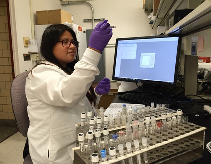A woman in a lab coat and glasses closely examines a test tube