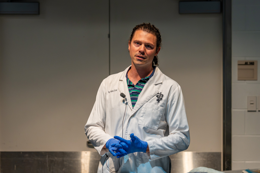 A man with long hair wearing a white coat stands in front of a stainless steel table. 