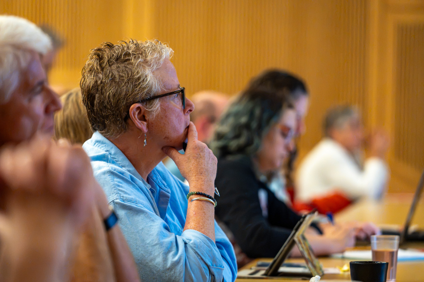 A woman with light hair and glasses takes in a lecture. 