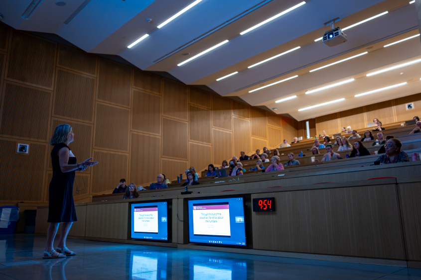 A woman in a blue dress stands in front of a crowd inside a lecture hall.