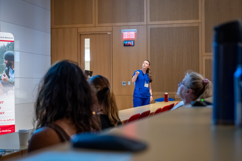 A woman in blue scrubs lectures to a large group. 