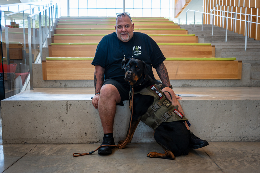 A man wearing all black sits with a large dog. 