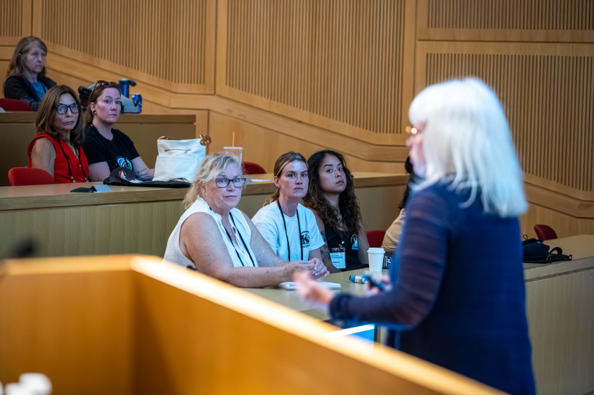 A woman with light hair lectures in front of a crowded room. 