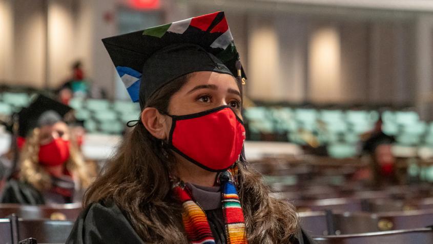 A veterinary student in a cap and gown with a mask on listens to the keynote speaker at the college's send-off ceremony