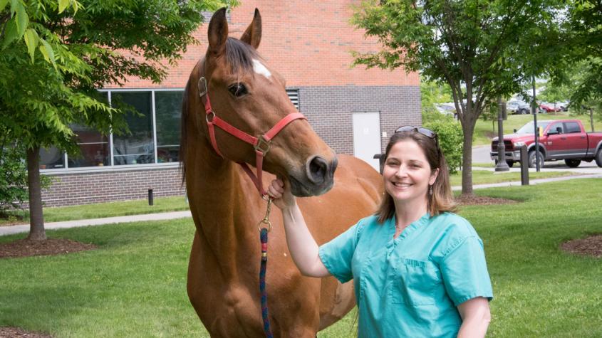 Michelle Delco in scrubs next to a brown horse outside of the college