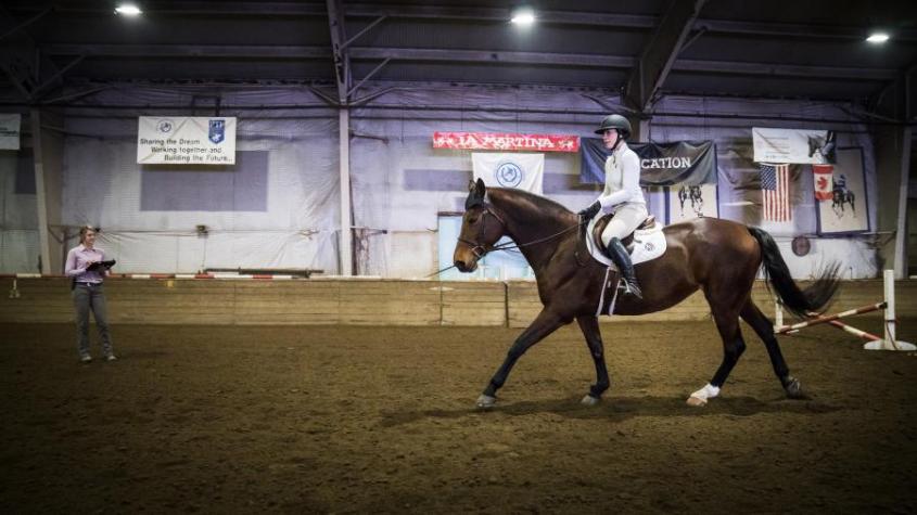 A rider on horseback in an arena as a veterinarian looks on. 