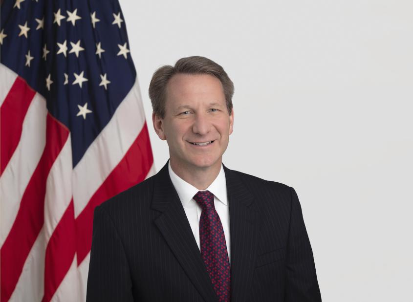A headshot of Dr. Normal Sharpless, wearing a suit and tie with an American flag backdrop