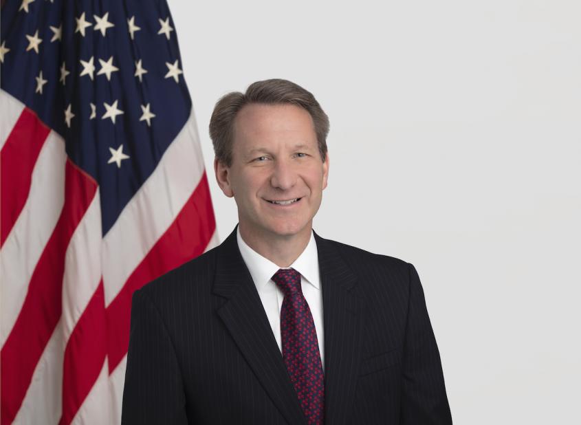 A headshot of Dr. Normal Sharpless, wearing a suit and tie with an American flag backdrop