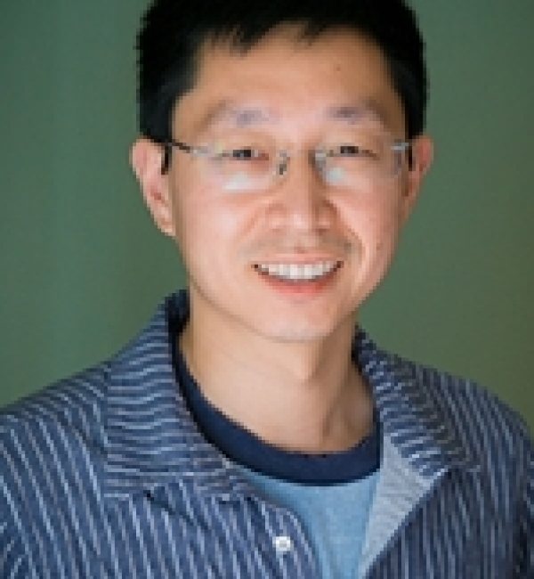 Chun Han in a green background with a blue and white striped collared shirt and wearing metallic thin-framed glasses.