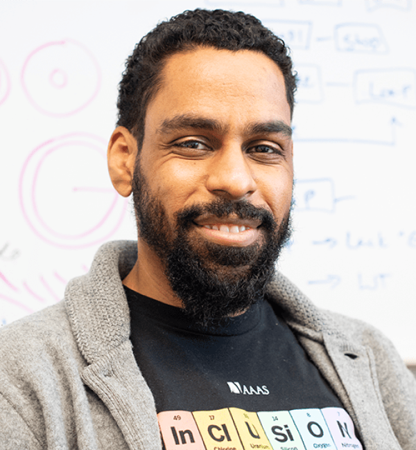 Karl Lewis wearing a chemistry novelty T-shirt and a grey zip-up hoodie in front of a white board with biochemical diagrams.