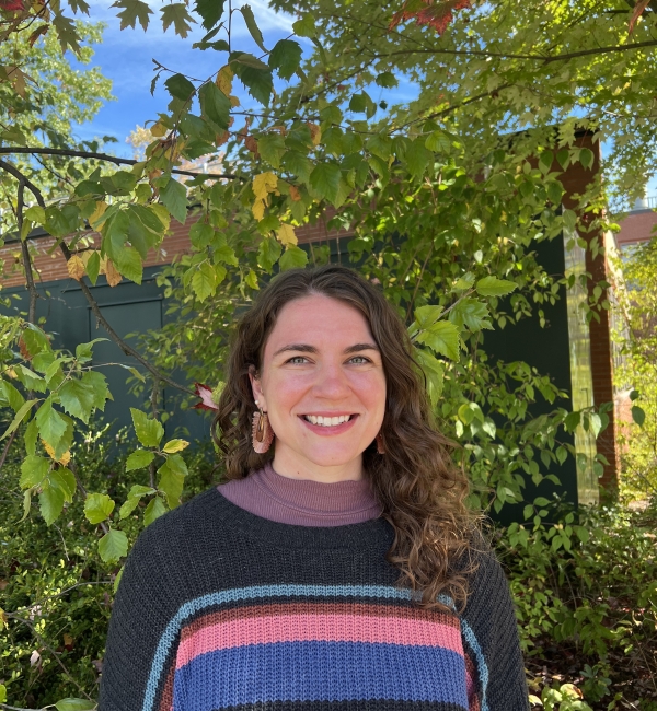 Sydney Womack wearing a knit sweater standing in front a building surrounded by green vegetation.