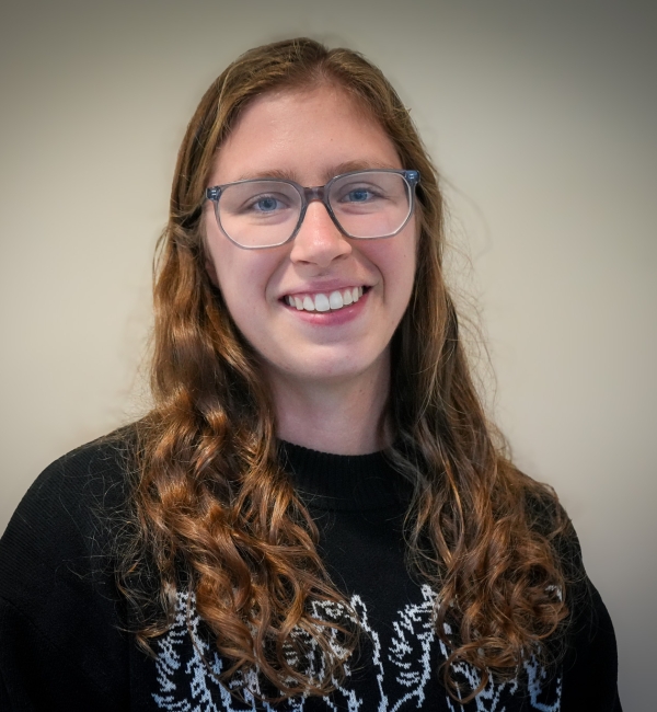 A woman with dark hair and glasses in front of a light wall. 