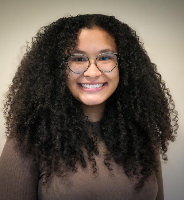 A woman with dark hair and glasses in front of a light wall. 