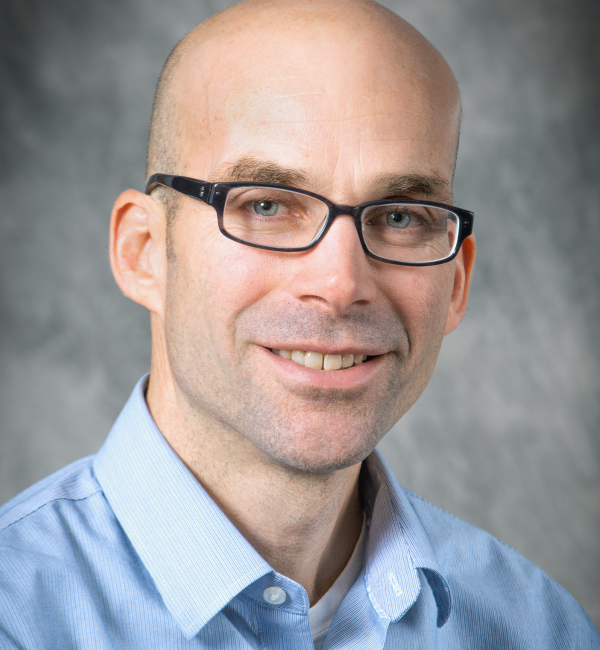 A man with blue yes and glasses against a gray background. 