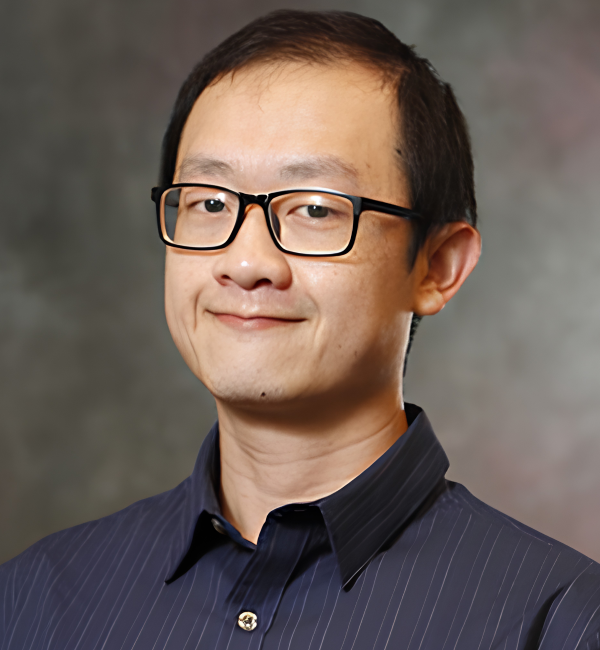 A man with glasses sits against a gray backdrop