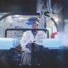 A male worker at the Cornell COVID Testing Lab, seen through stacks of samples; he is wearing a mask, a visor, and gloves