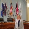 Krysten Schuler standing inside the House of Representatives, with flags and a giant desk behind her